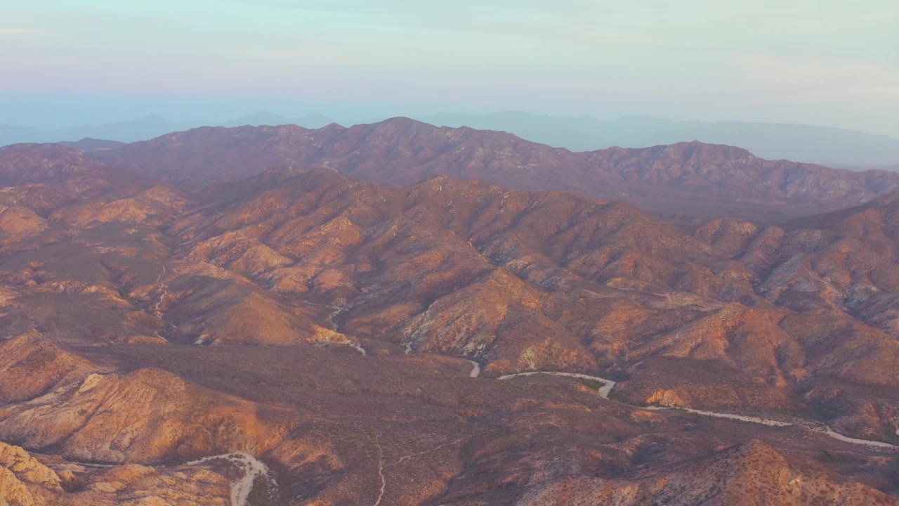 Wide aerial frame over Cabo Pulmo, showing layered brown ridges, a light road bending through the valley floor, distant mountains, and no visible people.