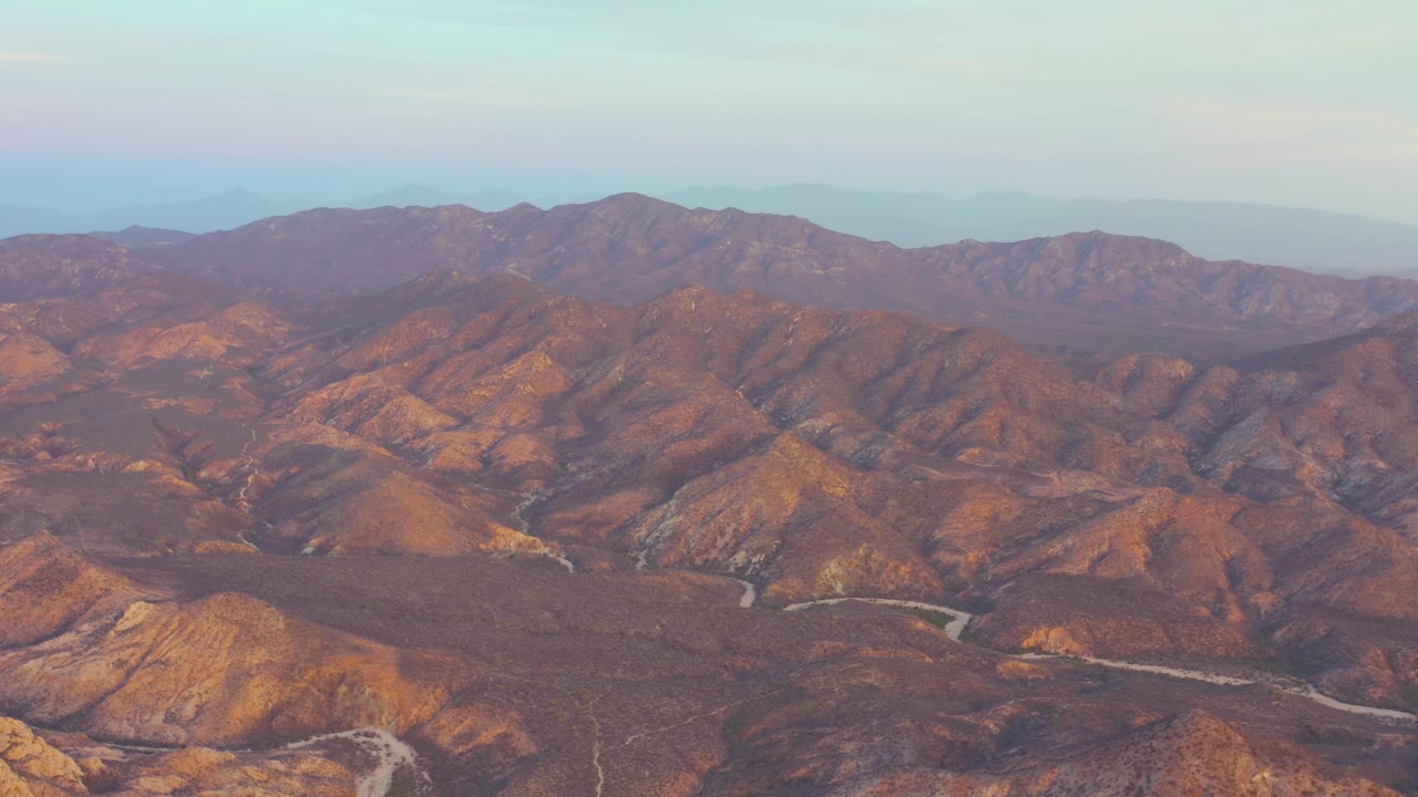 Aerial view in Cabo Pulmo with sunlit arid hills in the foreground, a curving road in the lower valley, darker ridges beyond, and no visible people.