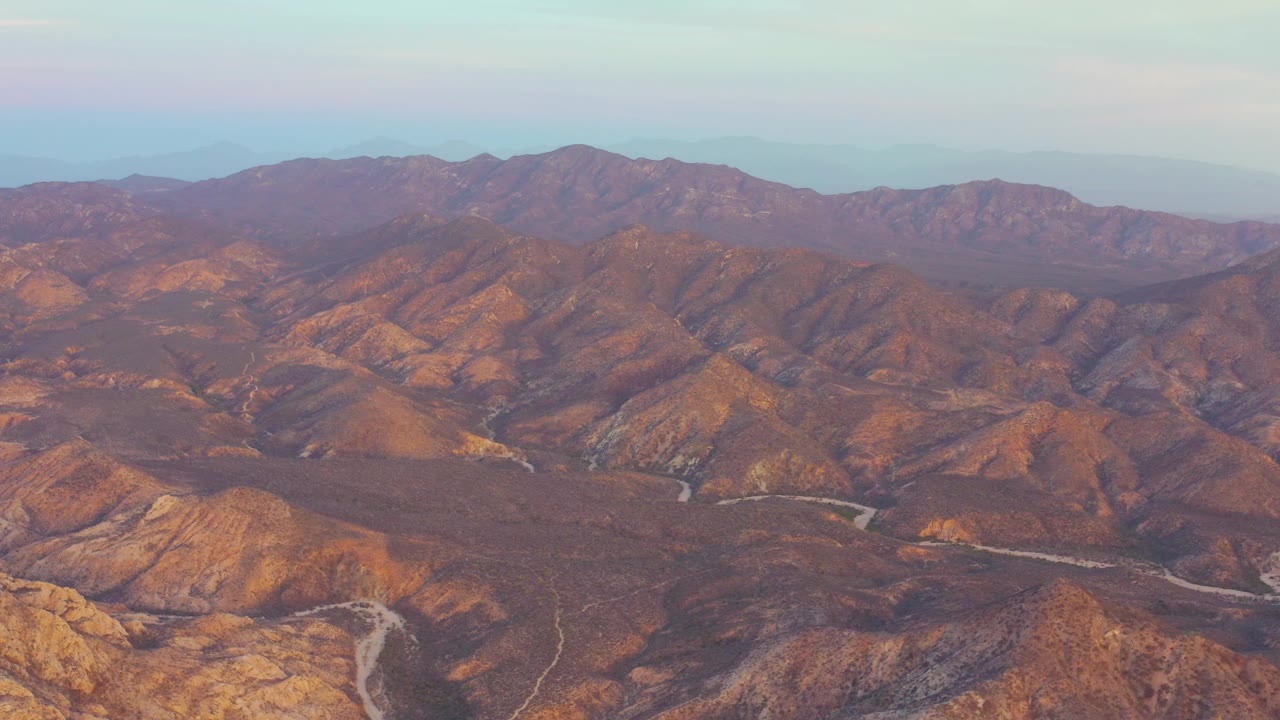 Aerial view near Cabo Pulmo, Baja California Sur showing orange-brown mountain ridges, pale road lines through the valley, distant hills, and no visible people.