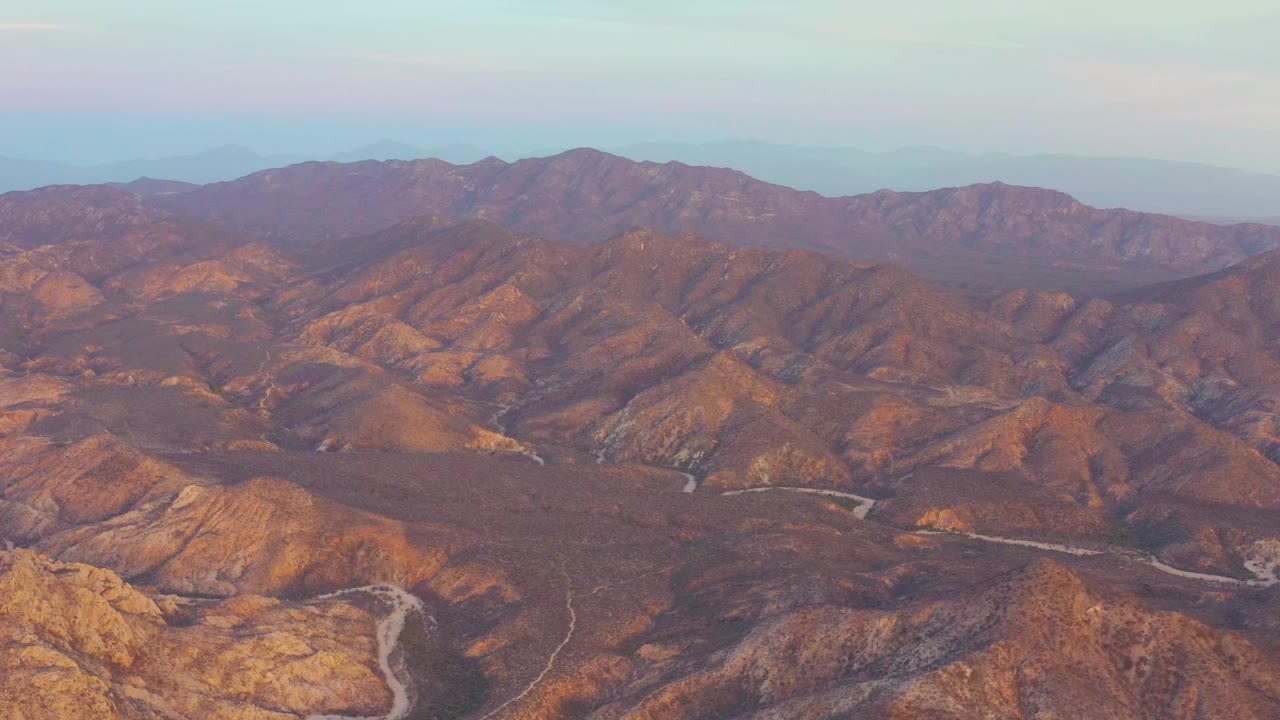 Aerial frame from Cabo Pulmo, Baja California Sur showing dry brown ridges, a pale winding road in the lower valley, distant mountains, and no visible people.
