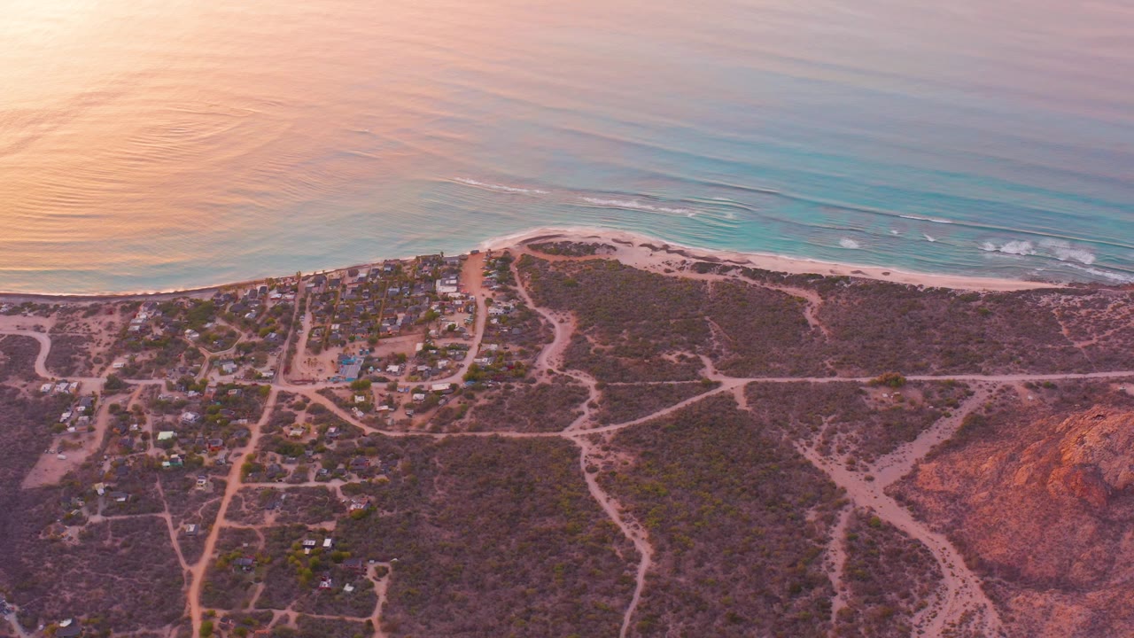 Cabo Pulmo coastline from above with the beach and turquoise ocean across the top of frame, small buildings and dirt roads below, warm pink light, and no visible people.