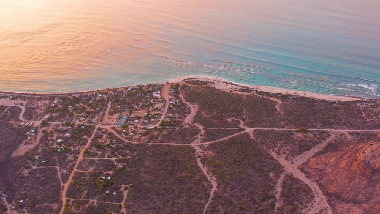 Aerial view over Cabo Pulmo with turquoise ocean, a narrow sandy beach, small residential roads, scattered homes, reddish dry terrain, and no visible people.