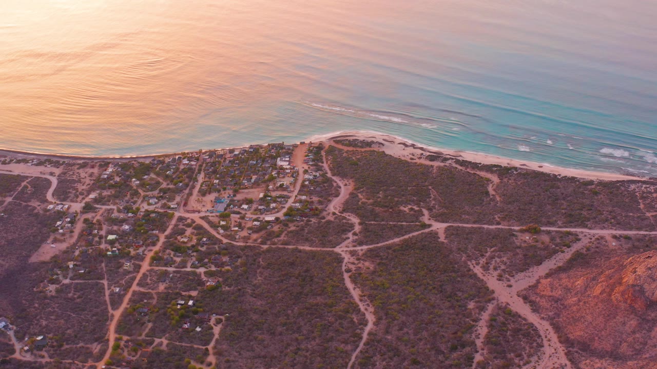 At 0:00, Cabo Pulmo appears from above with pink-toned ocean, a pale beach, branching dirt roads, dry scrubland, scattered small structures, and no visible people.