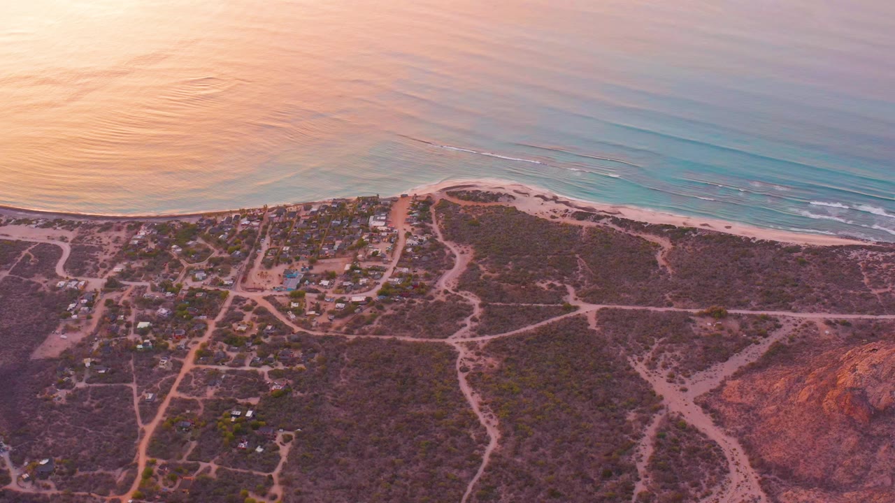 Aerial view one second into the Cabo Pulmo clip, with soft sunset color on the water, thin surf lines near shore, dirt tracks inland, scattered buildings, and no visible people.