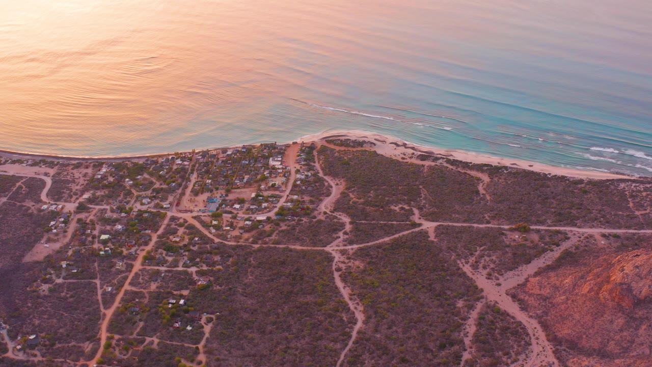 Cabo Pulmo aerial frame showing a broad band of turquoise water, narrow beach, inland scrubland crossed by pale roads, a cluster of buildings, and no visible people.