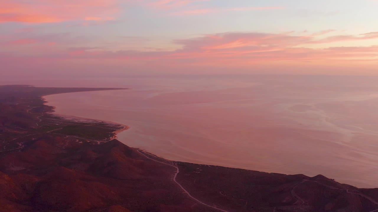 Cabo Pulmo aerial frame with a soft pink sky, broad calm water, dark coastal hills, a visible winding road, and no visible people.