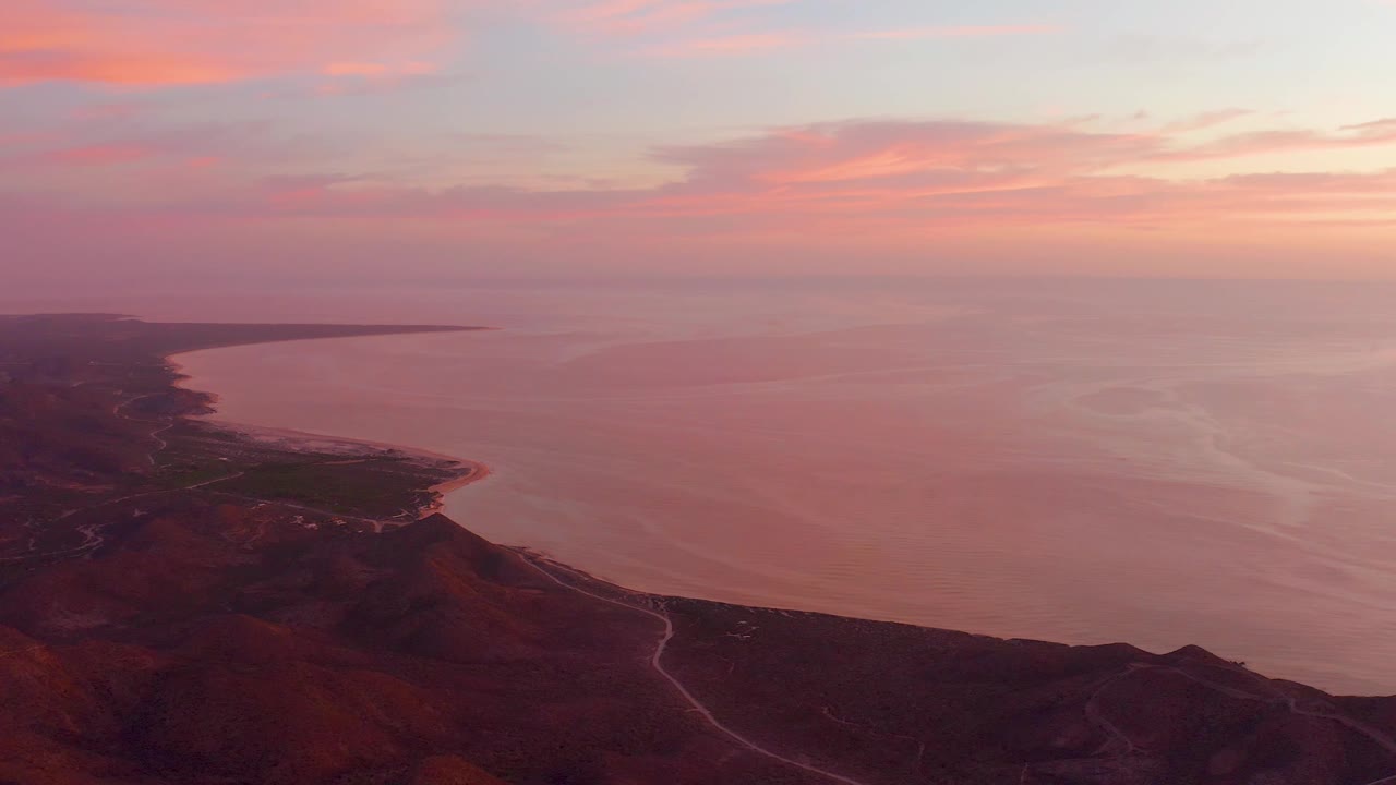 Aerial view over Cabo Pulmo coastline showing orange-pink clouds, calm sea, dark hills at the lower edge, a road trace, and no visible people.