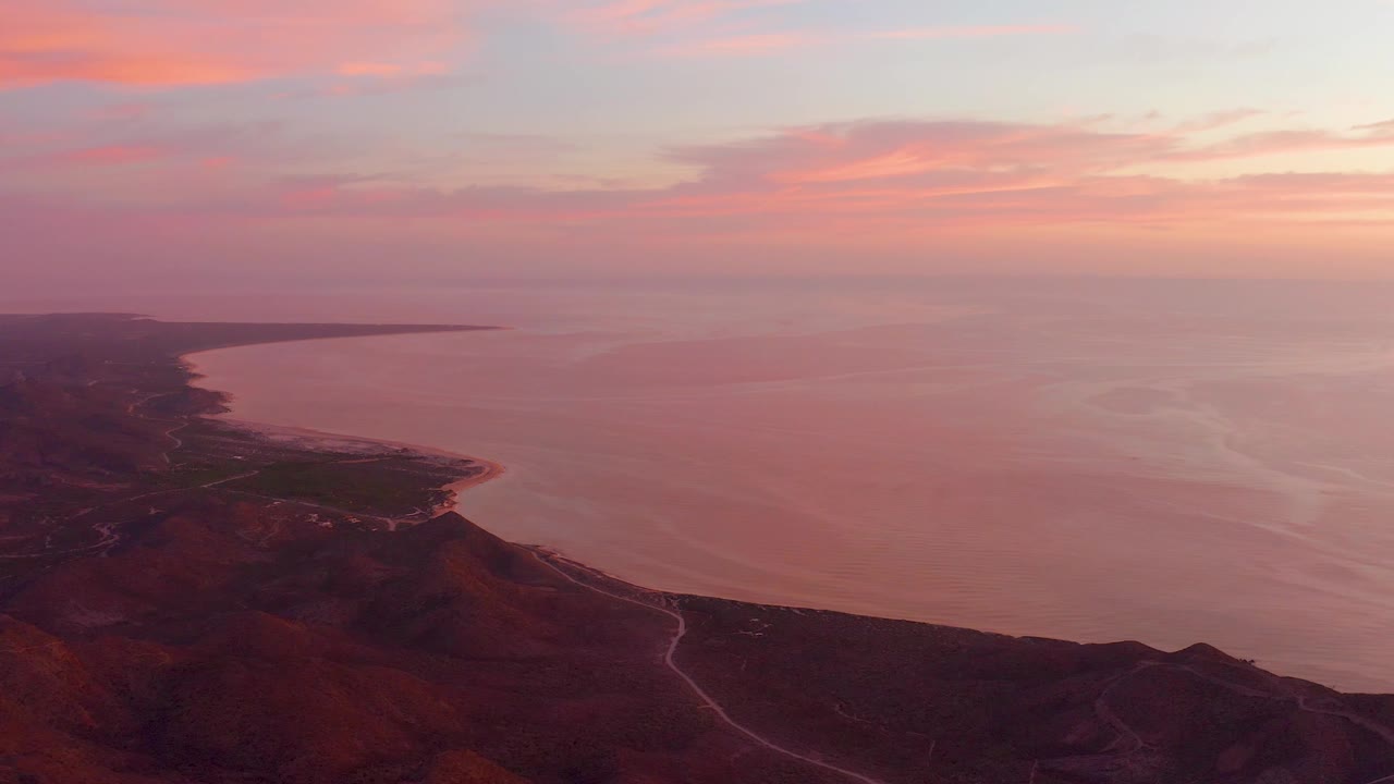 Cabo Pulmo sunset frame with the sea filling most of the view, darker hills and a winding road at the bottom, a long shoreline at left, and no visible people.