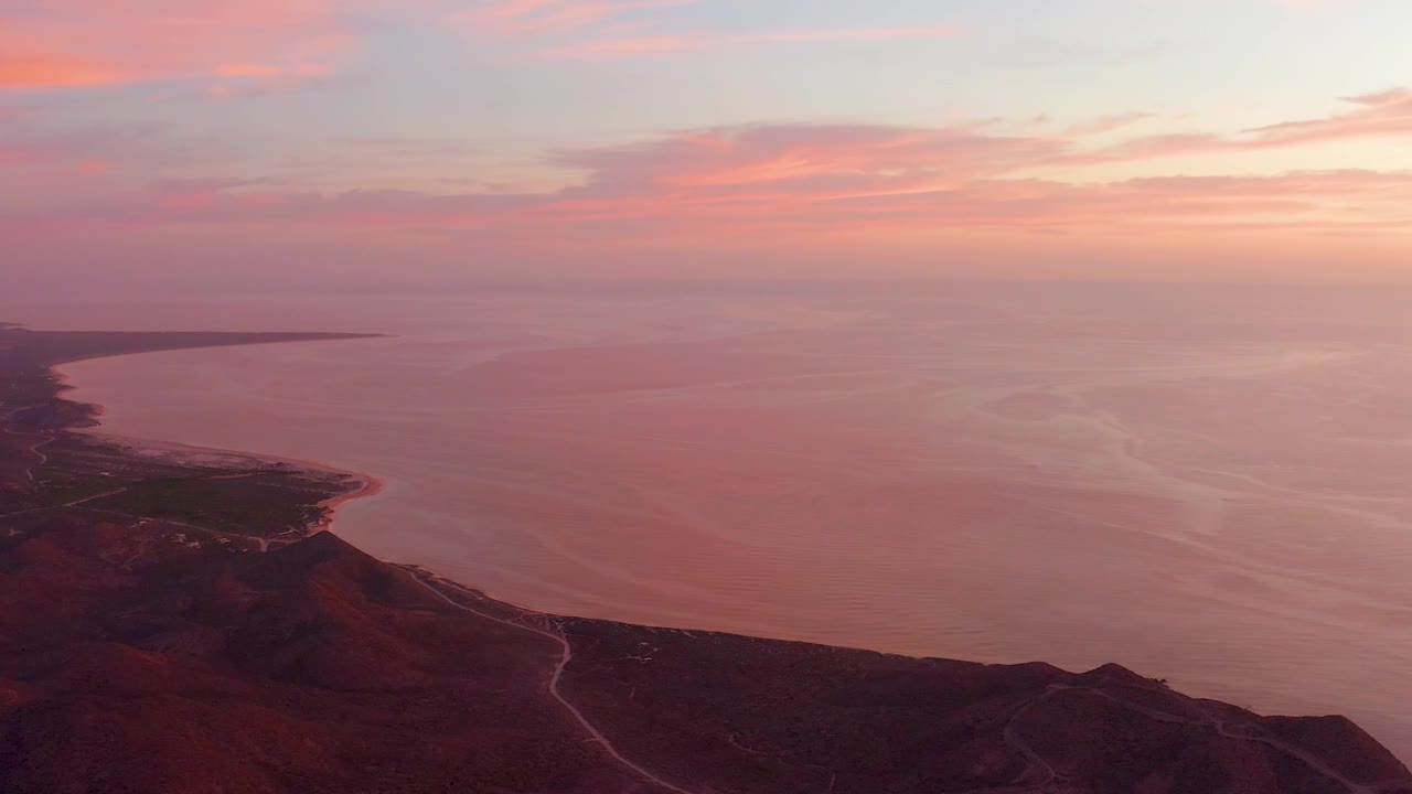 Cabo Pulmo sunset frame with the dark hill ridge occupying the lower edge, calm water stretching to the horizon, a small green patch near the shore, and no visible people.