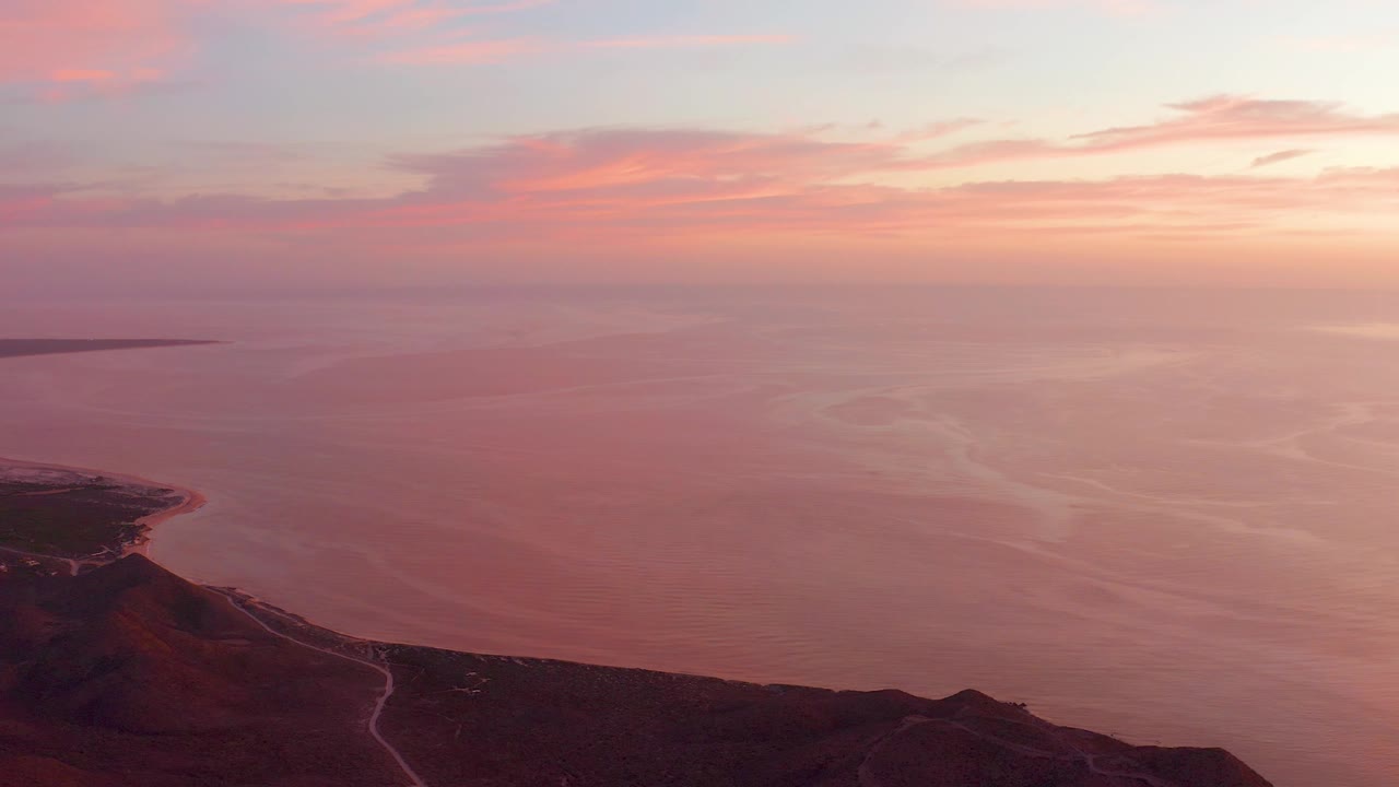 Cabo Pulmo sunset view over a pink ocean and dark coastal hillside, with a winding shore road in the lower frame and no visible people.