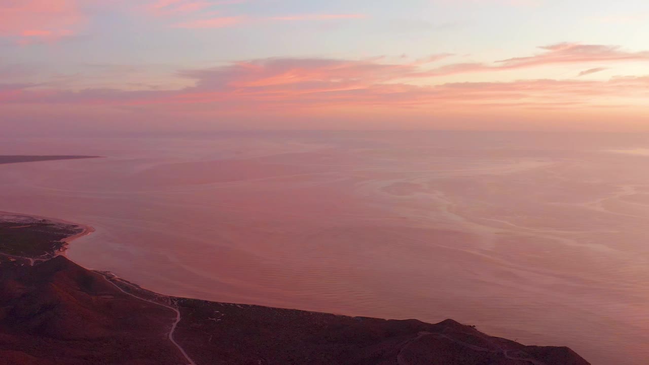 Cabo Pulmo ocean at sunset with pink clouds, a dark hillside road in the foreground, a distant low landform on the horizon, and no visible people.
