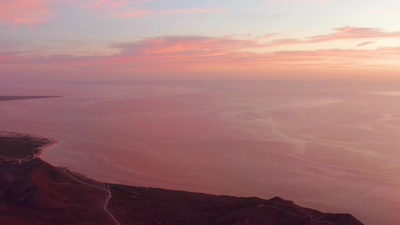 Cabo Pulmo coastline viewed from above with soft orange-pink sky, calm water, dark hillside foreground, a pale road trace, and no visible people.
