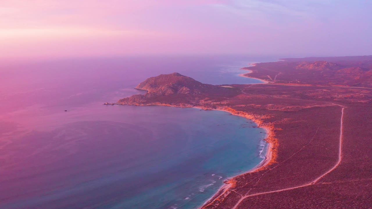 Wide aerial frame of Cabo Pulmo coastline showing the bay slightly farther across the frame, red-brown arid ground, a pale coastal road, distant headlands, and no visible people.