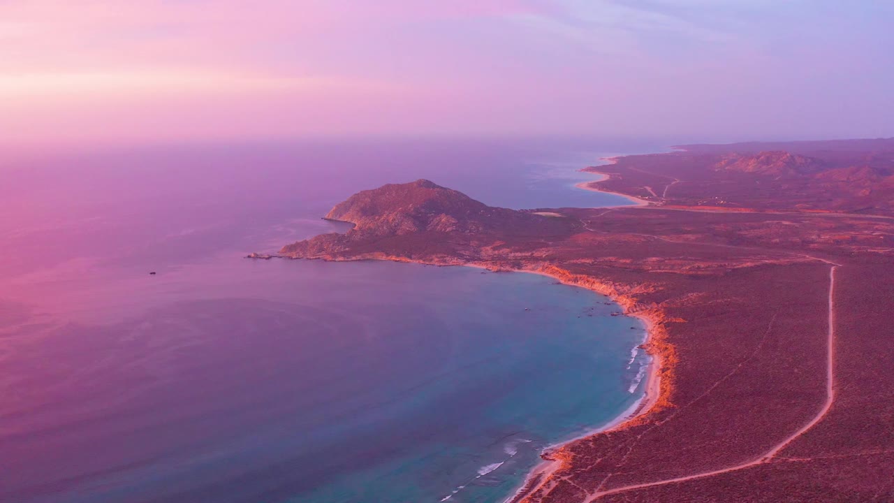 Cabo Pulmo coastline seen from above at 0:01, with the shoreline arc slightly farther across the frame, calm blue water below, arid land to the right, and no visible people.