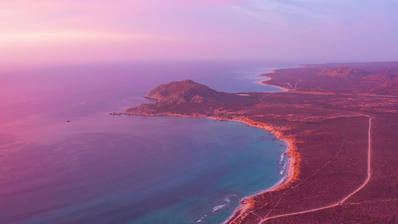 Aerial frame of Cabo Pulmo at 0:02 showing the blue bay in the lower frame, a narrow surf edge along the sandy beach, dry red-brown terrain, distant headland, and no visible people.