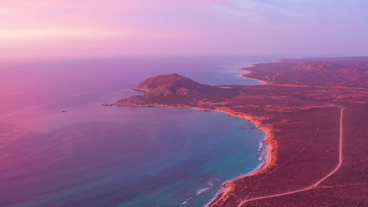 Cabo Pulmo aerial view at 0:03 with pink light over the ocean, a sweeping beach line, the coastal road forming a pale vertical curve inland, hills on the horizon, and no visible people.
