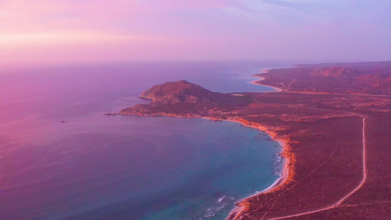 Cabo Pulmo coastline remains centered as the bay curves below the headland, with soft pink light over the water, orange-brown shore, a straight pale road inland, and no visible people.