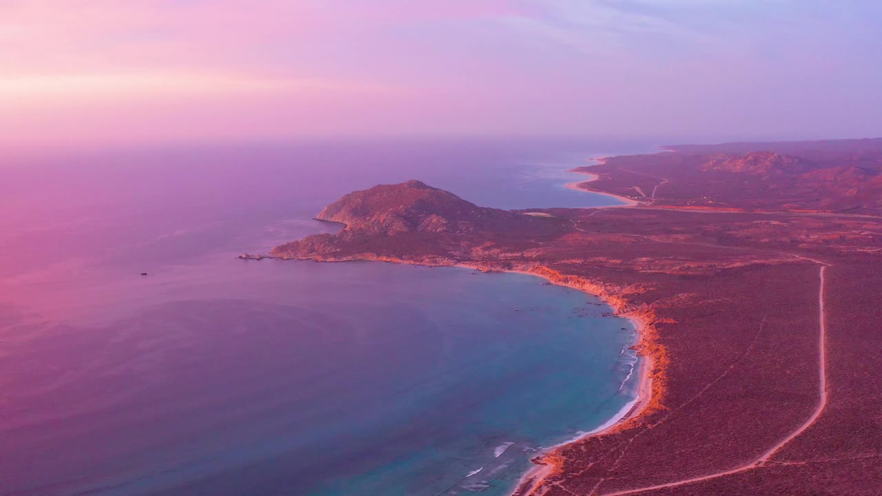 Mid-clip aerial view over Cabo Pulmo shows the headland slightly lower in frame, calm purple-toned water to the left, arid coastal plain to the right, a pale road, and no visible people.