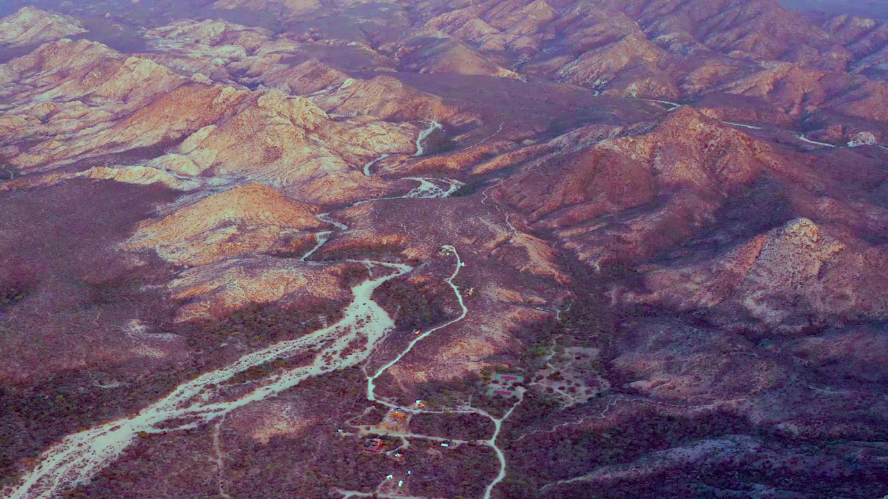 At 0:01, the aerial view holds on Cabo Pulmo's dry valley, where pale washes curve through brown and purple mountain terrain with no visible people.