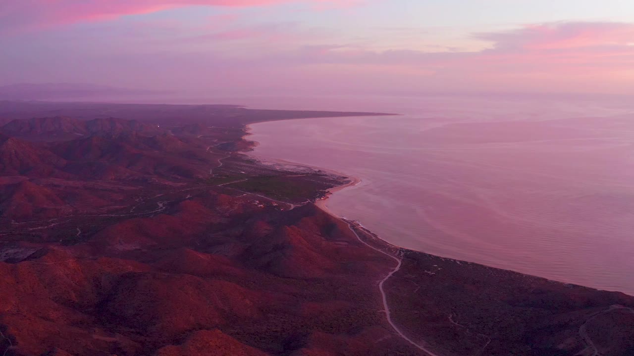 Cabo Pulmo aerial view with red ridges across the lower frame, the shoreline curving into calm water, a thin road tracing the coast, pink cloud bands near the top, and no visible people.