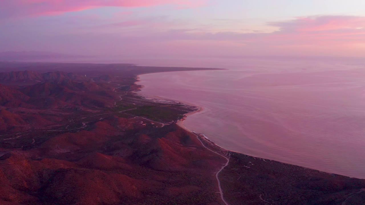 Aerial dusk view over Cabo Pulmo, Baja California Sur, with red ridges in the foreground, a narrow road near the shore, calm ocean to the right, and no visible people.