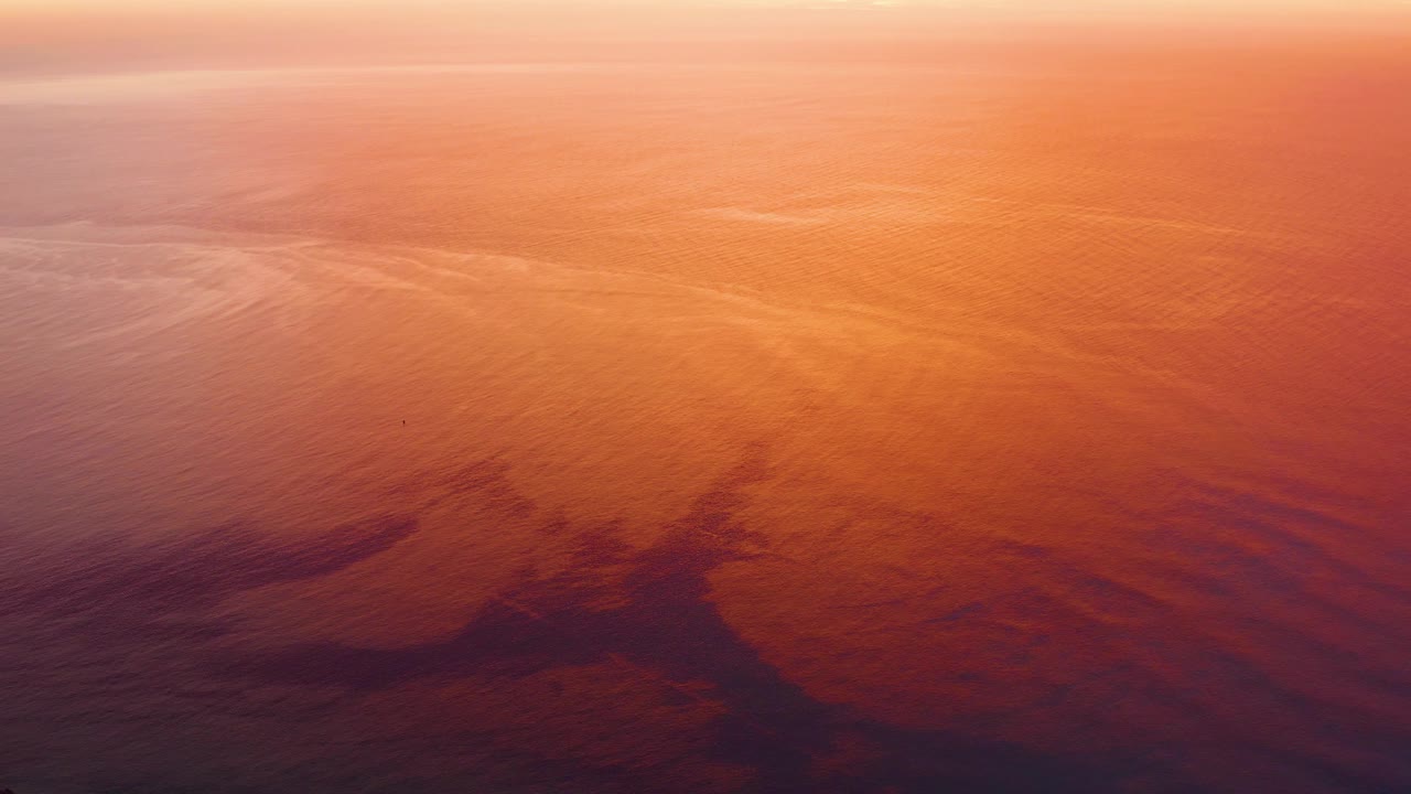 Cabo Pulmo ocean surface at 0:02 with orange sunset color, fine wave texture, faint pale streaks across the upper water, and no visible people.