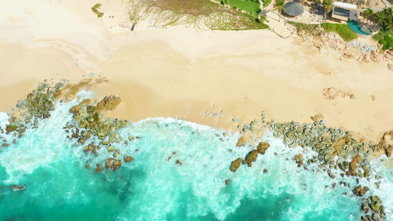 Aerial frame of Marquis Los Cabos shoreline with white foam widening over the center rocks, pale sand above, shoreline vegetation, structures, and no visible people.