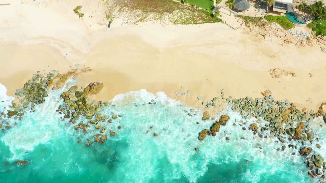 Aerial Marquis Los Cabos shoreline frame with foamy surf pushing diagonally along rocks, empty sand filling the upper half, sparse vegetation, structures, and no visible people.