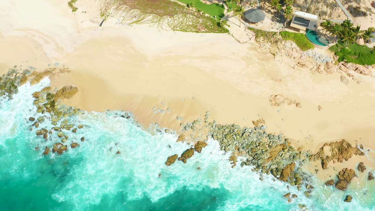 At 0:01, the overhead frame shows foam spreading farther across the central rocks, tan sand above, and a small pool and building near the upper edge at Marquis Los Cabos, with no visible people.