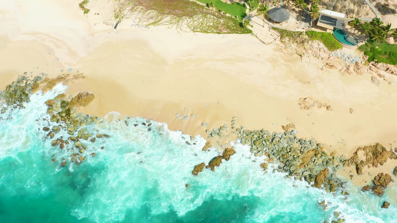 At 0:03, the surf line sits lower across the frame while foam pushes toward the rocks and the sandy beach opens broadly above it at Marquis Los Cabos, with no visible people.