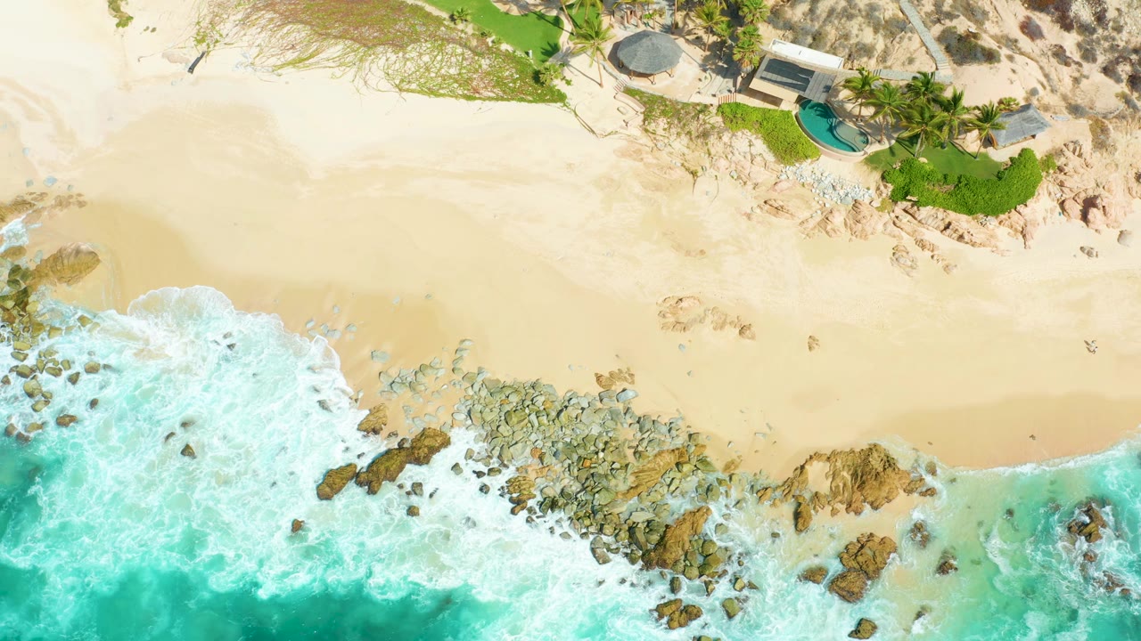 Aerial frame at Marquis Los Cabos with foamy turquoise water wrapping around shoreline rocks, a broad band of sand, resort landscaping at the top, and no visible people.