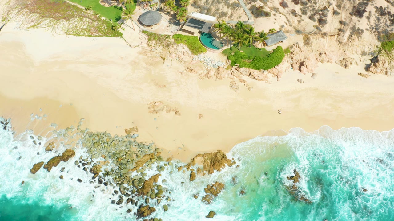 Overhead frame at Marquis Los Cabos shows turquoise surf breaking over rocks below pale sand, with a small building, green lawn, palms, and pool near the dunes, and a tiny non-recognizable person-like figure on the beach.