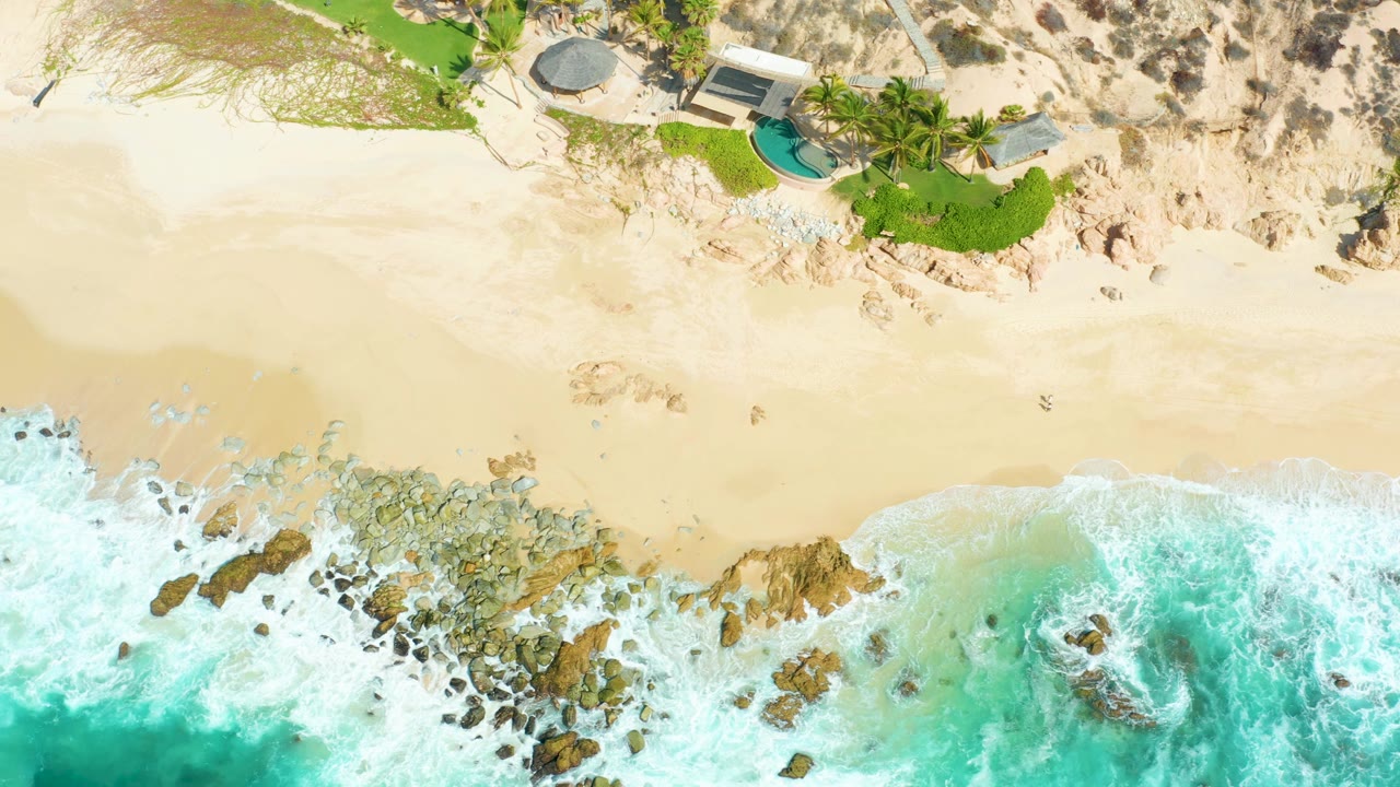 At 0:02, white surf pushes farther across the rocky lower shore while a broad band of pale sand separates the water from the green landscaped area and pool at Marquis Los Cabos.