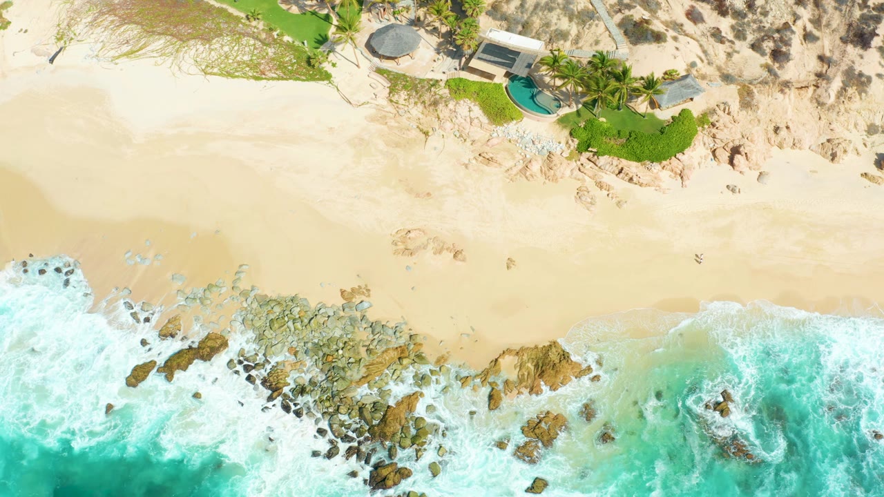 Aerial beach frame with waves breaking on rocks and a small pool near the dunes.