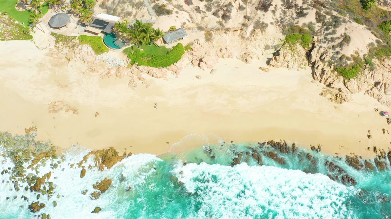 Overhead view at Marquis Los Cabos showing turquoise surf breaking over rocks below pale sand, with cliffside homes, pools, palms, grass, and no visible people.
