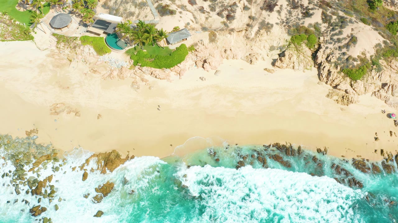 Aerial frame at Marquis Los Cabos with a broad band of pale sand between foamy turquoise water and cliffside homes with green lawn and pools, with no visible people.