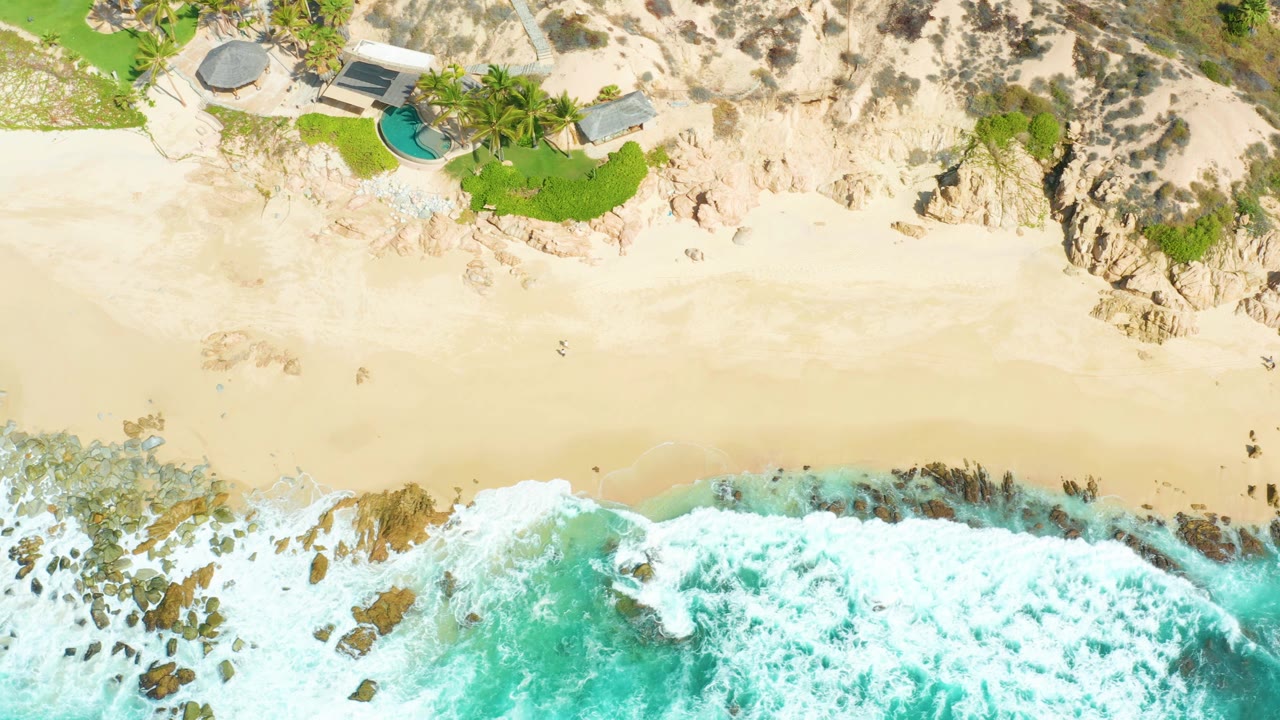 Drone view at Marquis Los Cabos where a fresh wave line reaches the lower beach, rocks cut through the surf, and homes with pools sit above the sand, with no visible people.
