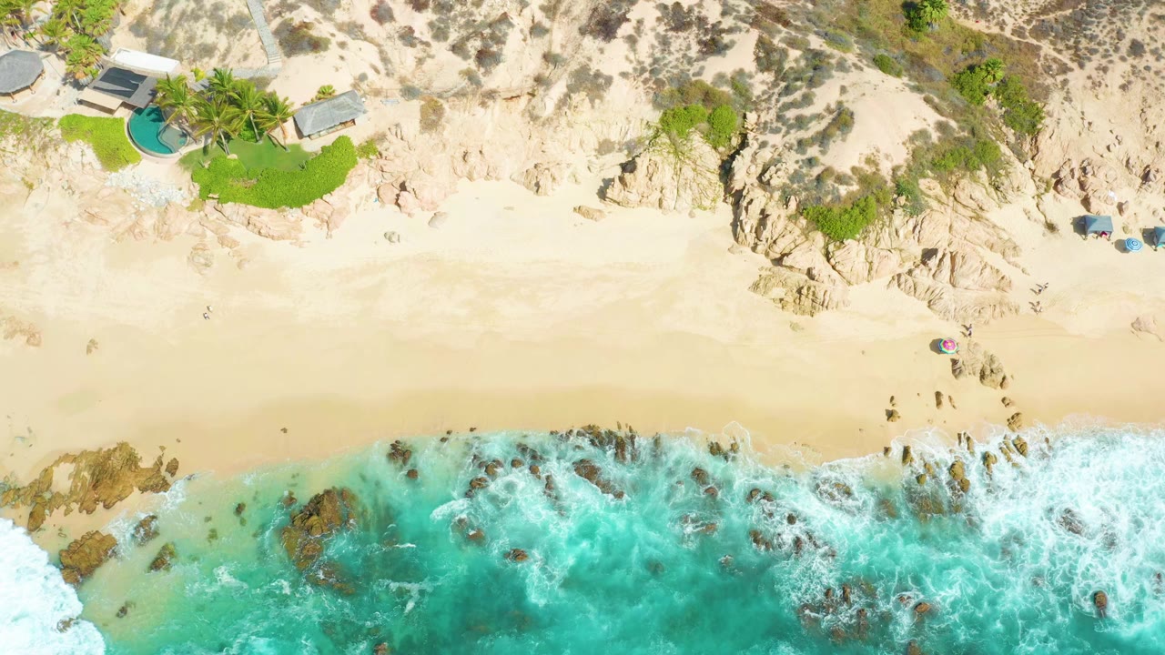 Aerial beach frame with waves breaking over rocks below cliffs and resort grounds.