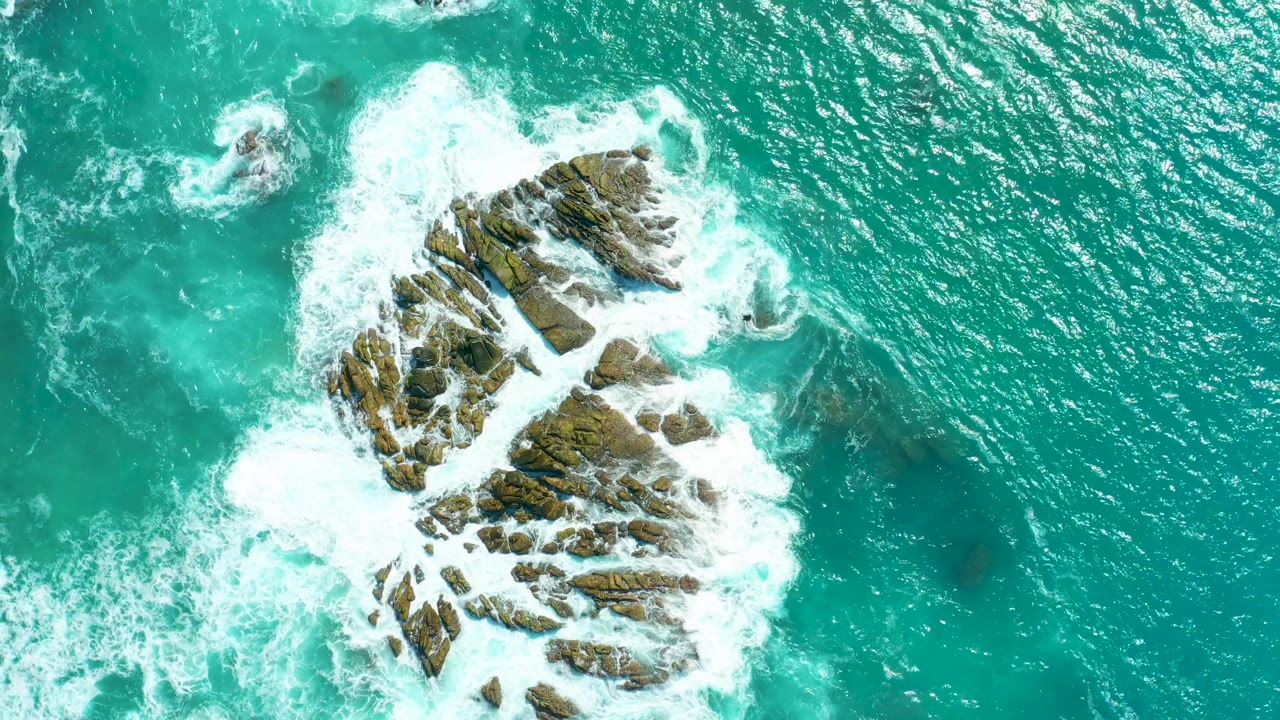 Aerial frame from Marquis Los Cabos showing a fresh band of white foam spreading across the upper rocks and teal water, with no visible people.