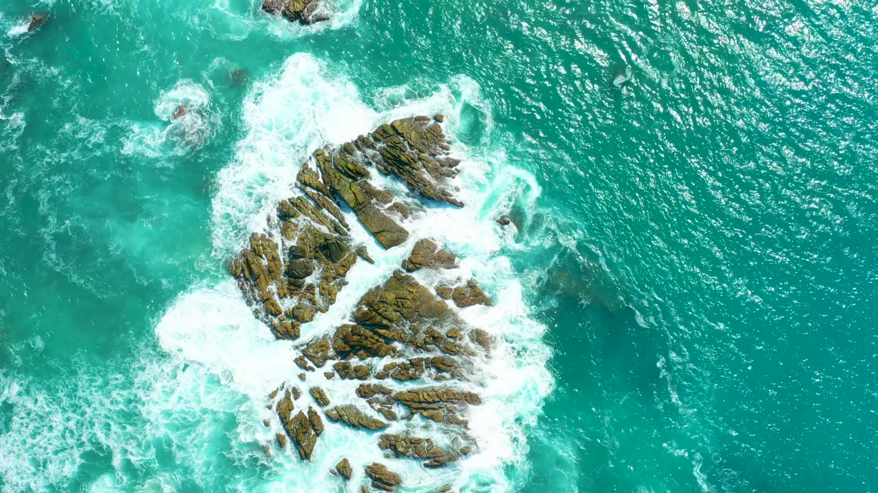 Aerial view at Marquis Los Cabos with a larger foam patch striking the lower edge of the rocky outcrop and teal water surrounding it, with no visible people.
