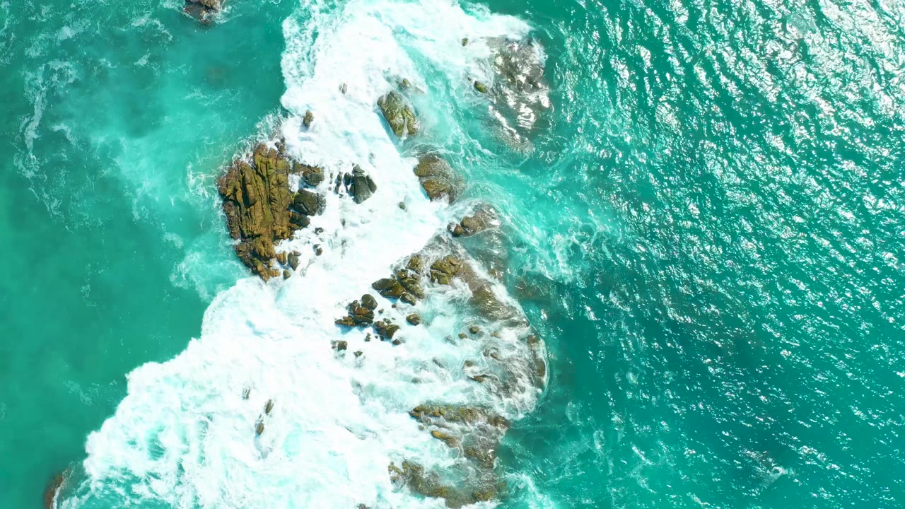 Aerial view at Marquis Los Cabos showing turquoise water and white surf wrapping around scattered brown rocks, with no visible people.
