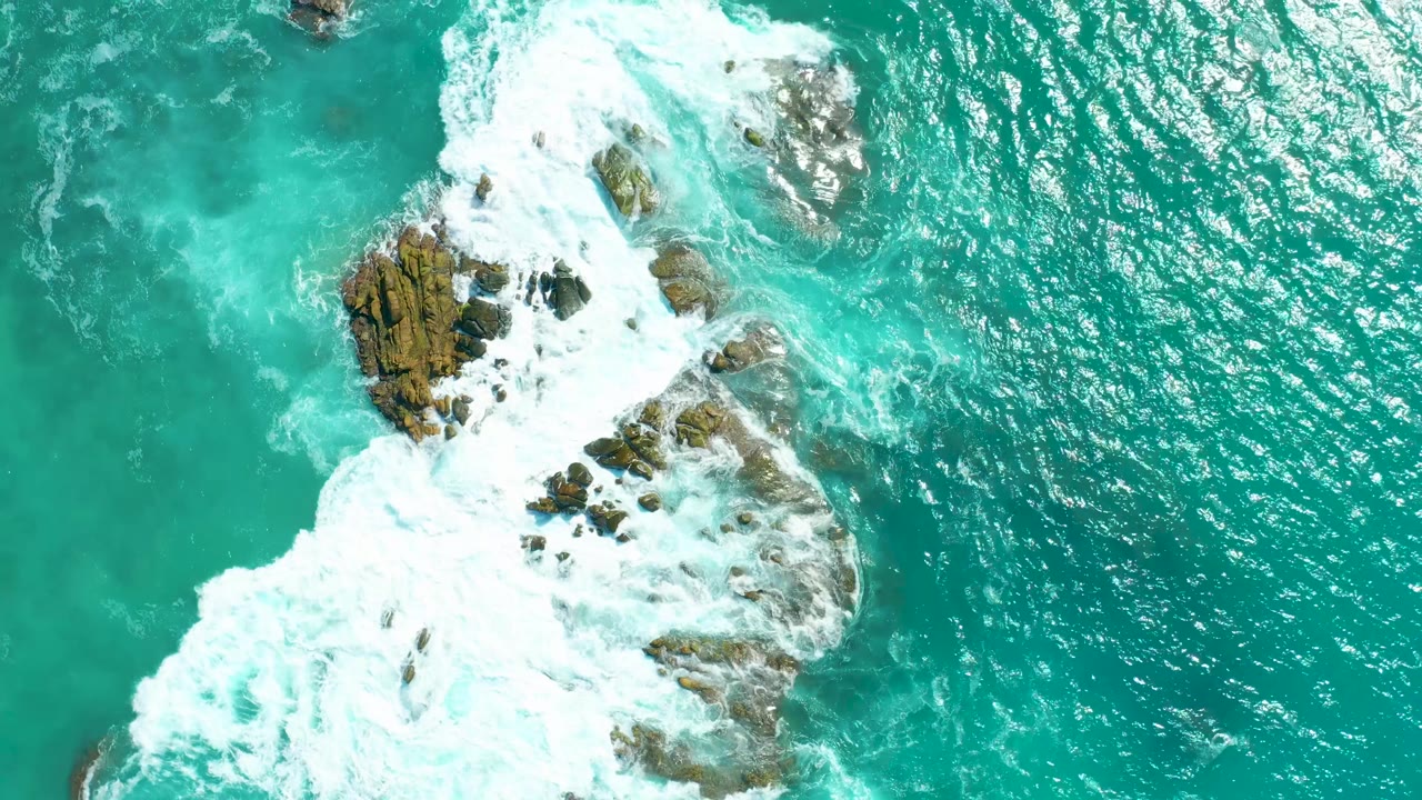 Whitewater spreads over rocky outcrops in bright turquoise sea.