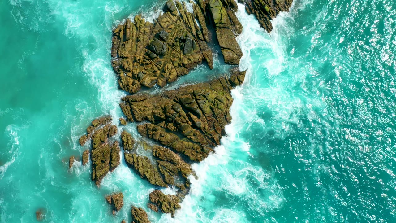 Overhead Marquis Los Cabos view of a large rock outcrop on the left, smaller rocks below it, and white surf curling through turquoise water, with no visible people.