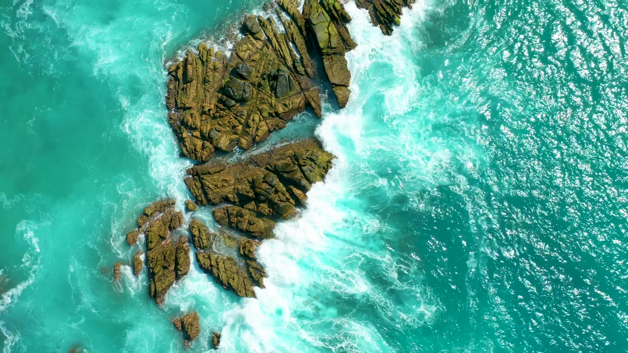 Top-down frame at Marquis Los Cabos showing the rock group shifted lower in view as white surf breaks along its right edge and open turquoise water fills the frame, with no visible people.