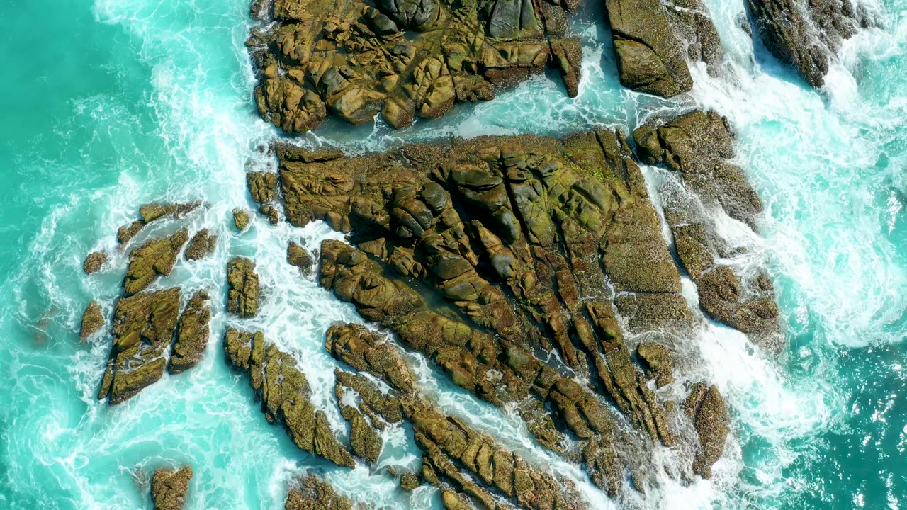 Aerial view at Marquis Los Cabos with white surf pushing through narrow channels between brown rock formations and turquoise water, with no visible people.