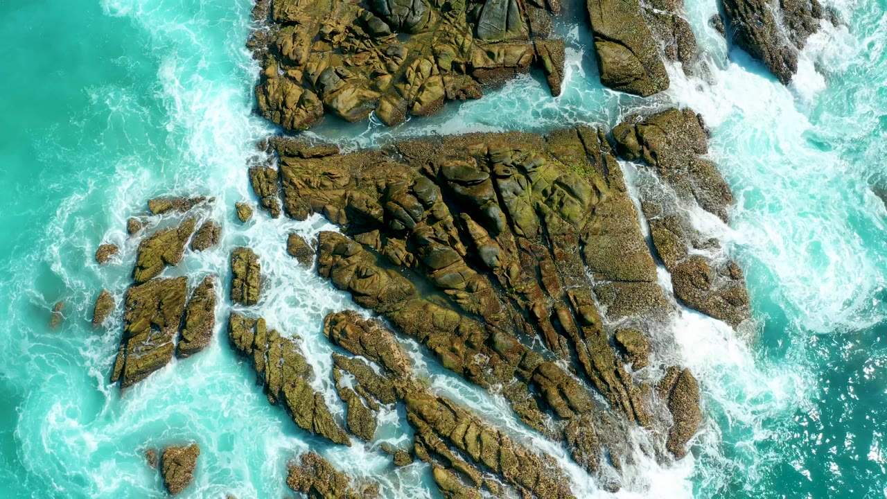 Top-down coastal view in Baja California Sur where foamy surf breaks against clustered rock outcrops in clear turquoise water, with no visible people.
