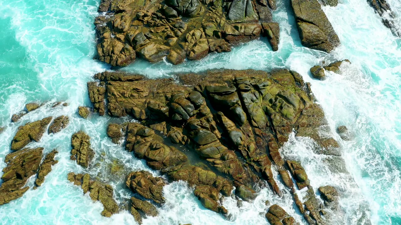 Foam spreads farther across the right side of the Marquis Los Cabos rock formations as turquoise water fills the left side, with no visible people.