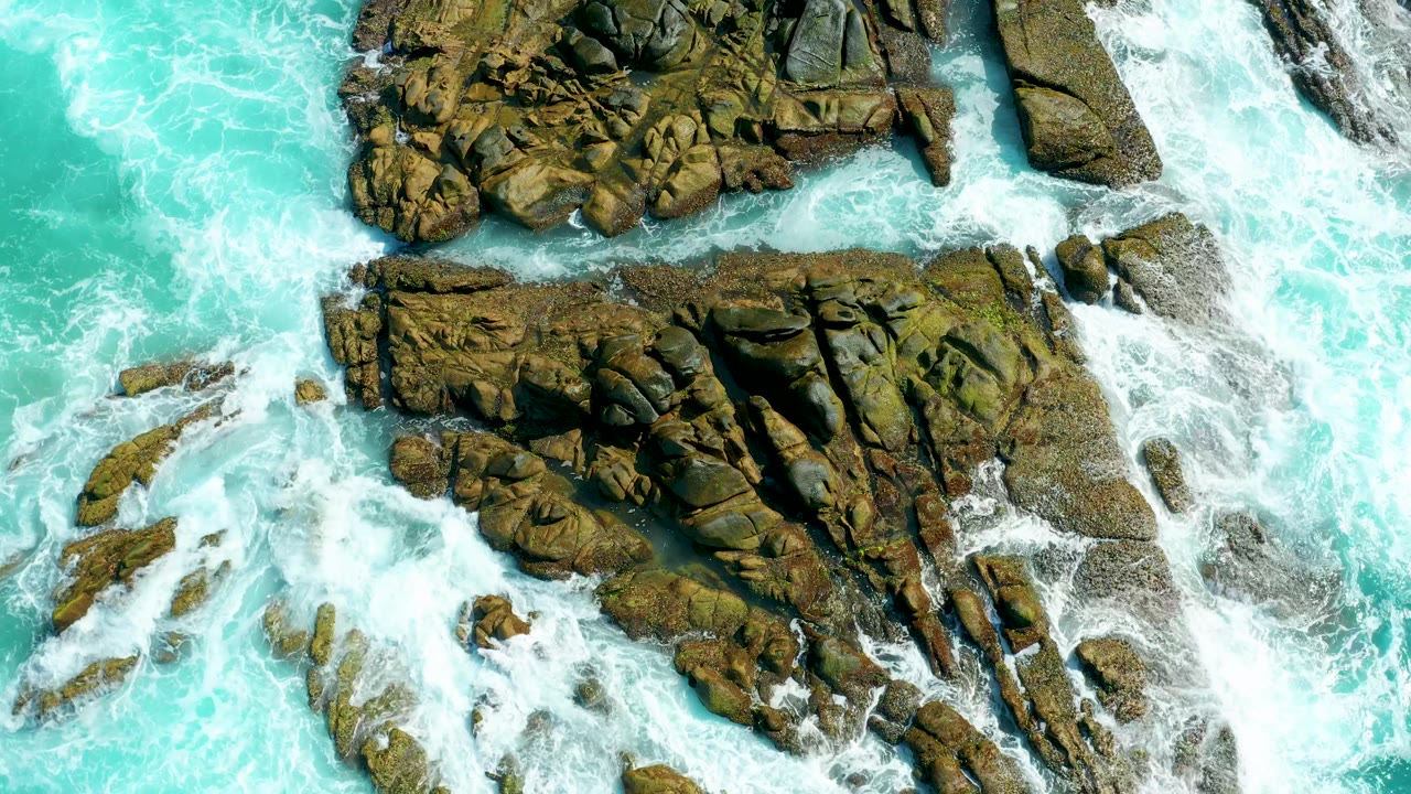 The rock formation remains centered from above as bright turquoise surf pushes around its left and lower edges at Marquis Los Cabos, with no visible people.