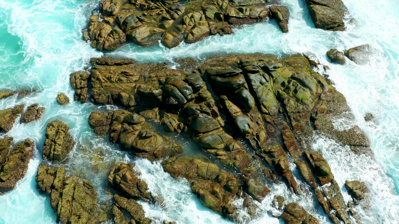 Overhead frame from Marquis Los Cabos showing turquoise water and white foam wrapping around jagged brown rocks, with no visible people.