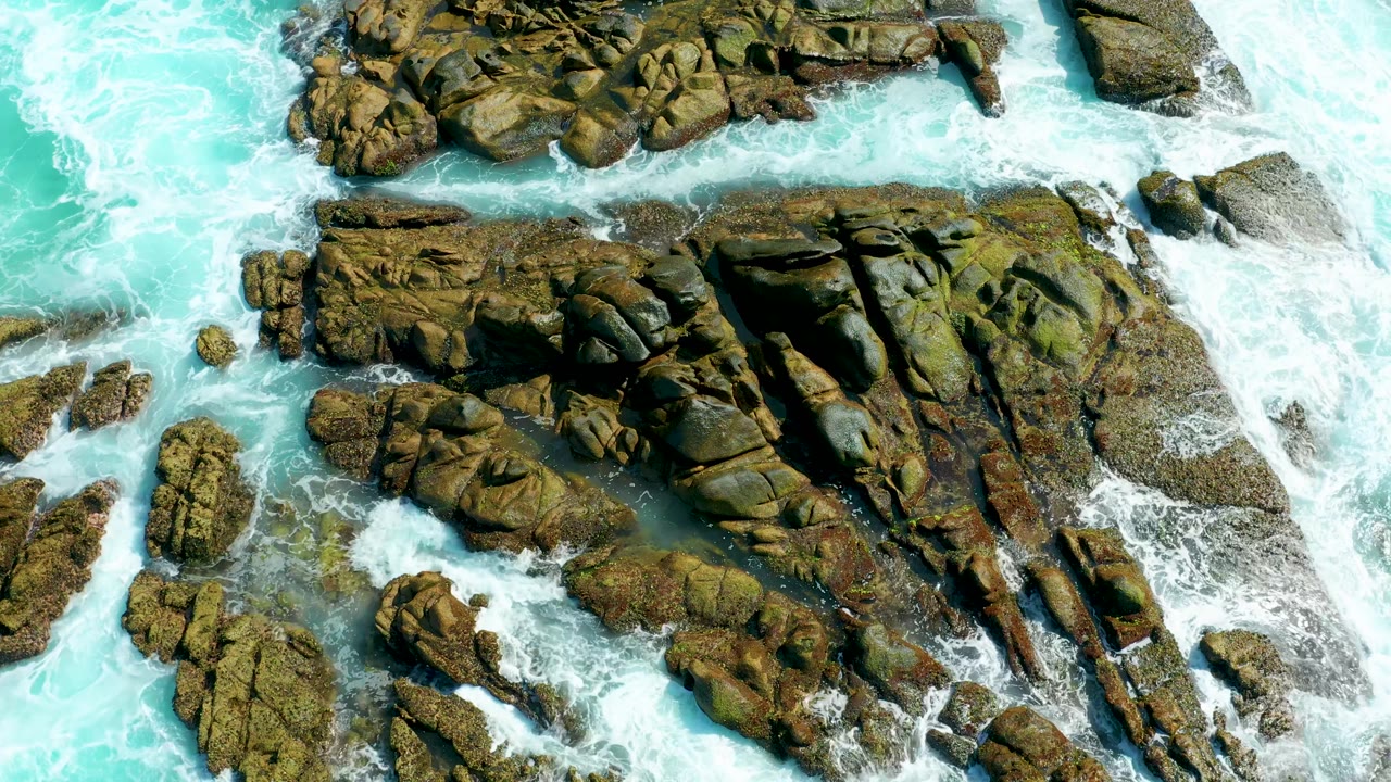 At 0:02, breaking surf pushes through narrow gaps beside the large central rock formation at Marquis Los Cabos, with no visible people.